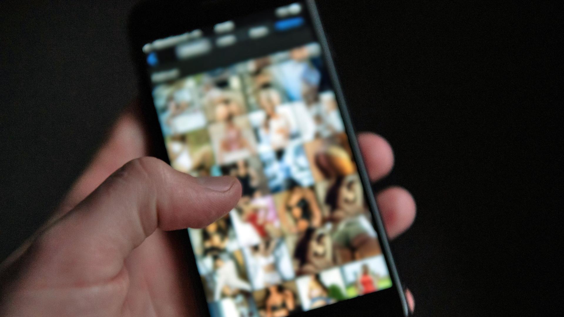 Eine Männerhand hält ein Smartphone, auf dem Bilder einer Pornografie-Internetseite zu sehen sind. 