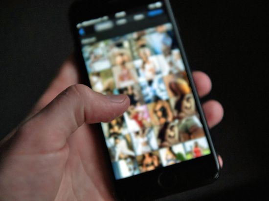 Eine Männerhand hält ein Smartphone, auf dem Bilder einer Pornografie-Internetseite zu sehen sind. 