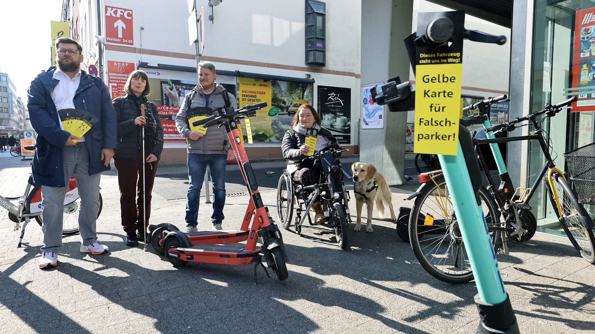 Vier Menschen halten eine schwarz-gelbe Karte in der Hand. Diese Karte mit der Aufschrift „Gelbe Karte für Falschparker!“ hängt auch am Lenker eines E-Scooters.