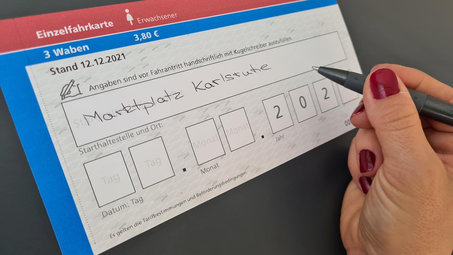 Fahrkarte, in den eine Hand mit einem Kuli „Marktplatz Karlsruhe“ einträgt.