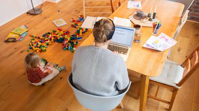 Ein zweijähriges Kind spielt im Wohnzimmer, während seine Mutter Zuhause im Homeoffice an einem Laptop arbeitet. 