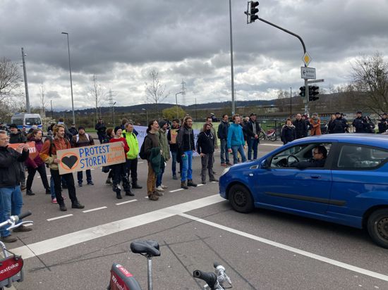 Mehrere Menschen stehen mit Schildern auf einer Kreuzung und hindern Autos am Weiterfahren. 