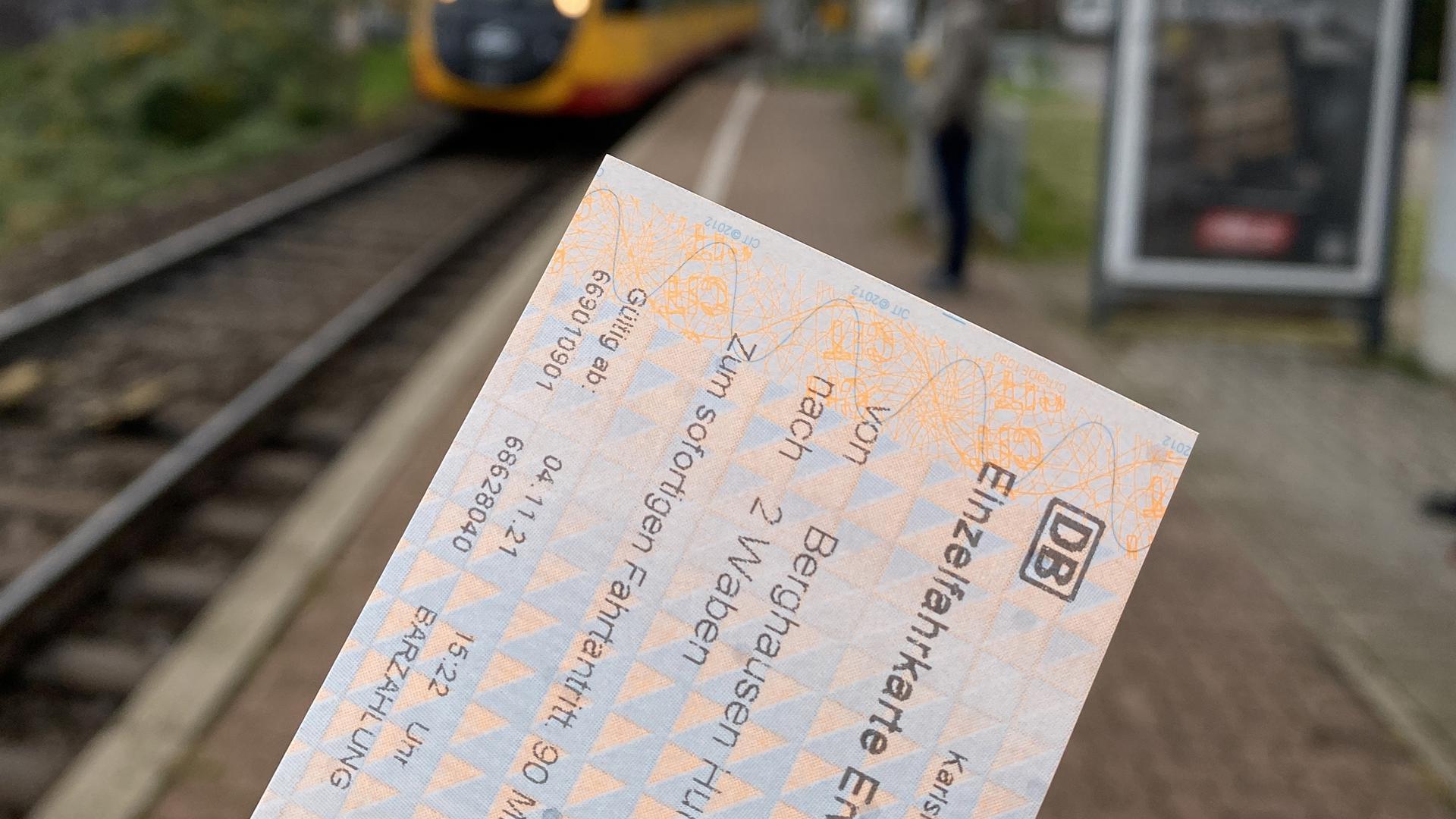 Eine Hand zeigt im Vordergrund eine KVV-Fahrkarte, im Hintergrund fährt eine Bahn an einer Haltestelle ein.