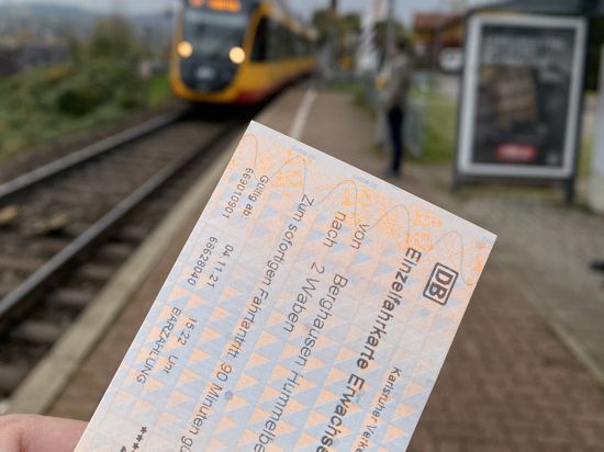 Eine Hand zeigt im Vordergrund eine KVV-Fahrkarte, im Hintergrund fährt eine Bahn an einer Haltestelle ein.