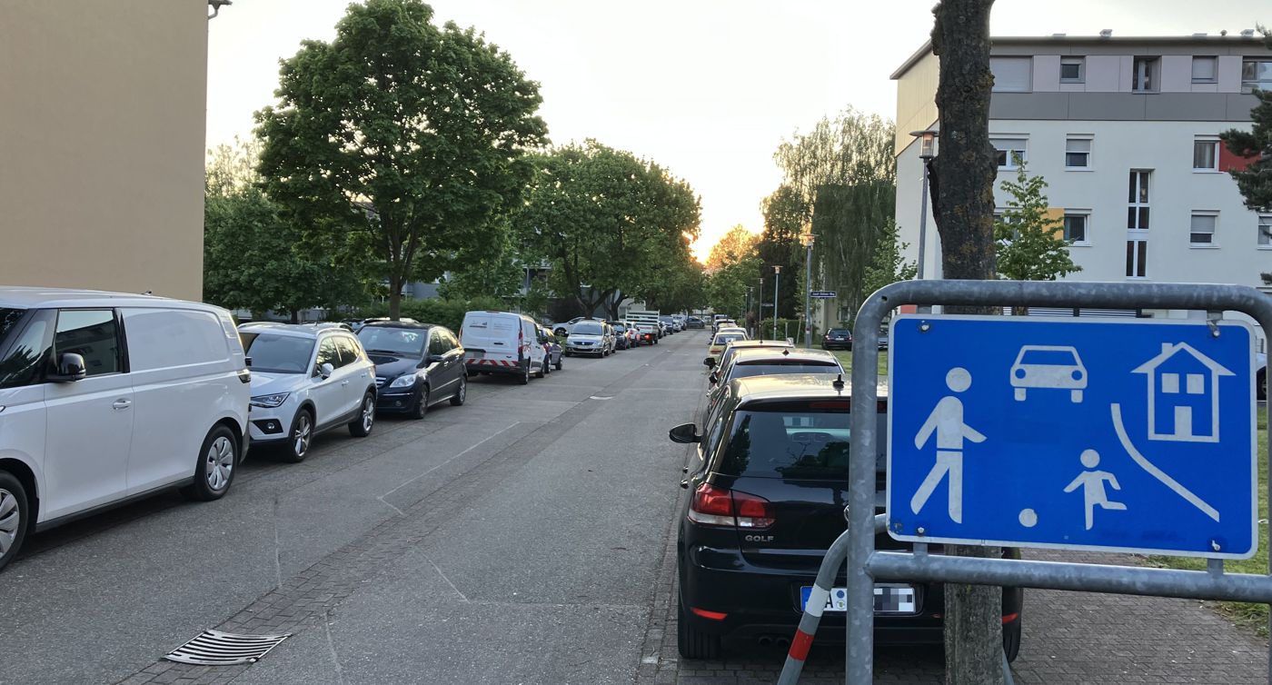 Josef-Stöhrer-Weg: Eine Straße, rechts und links Autos, vorne ein Spielstraßenschild. Links ein Wohnblock.