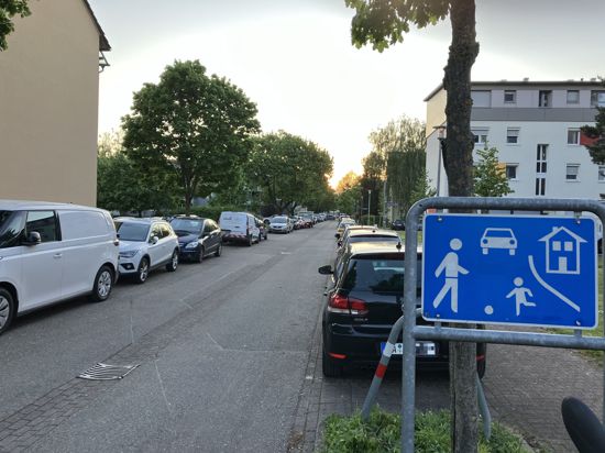 Josef-Stöhrer-Weg: Eine Straße, rechts und links Autos, vorne ein Spielstraßenschild. Links ein Wohnblock.