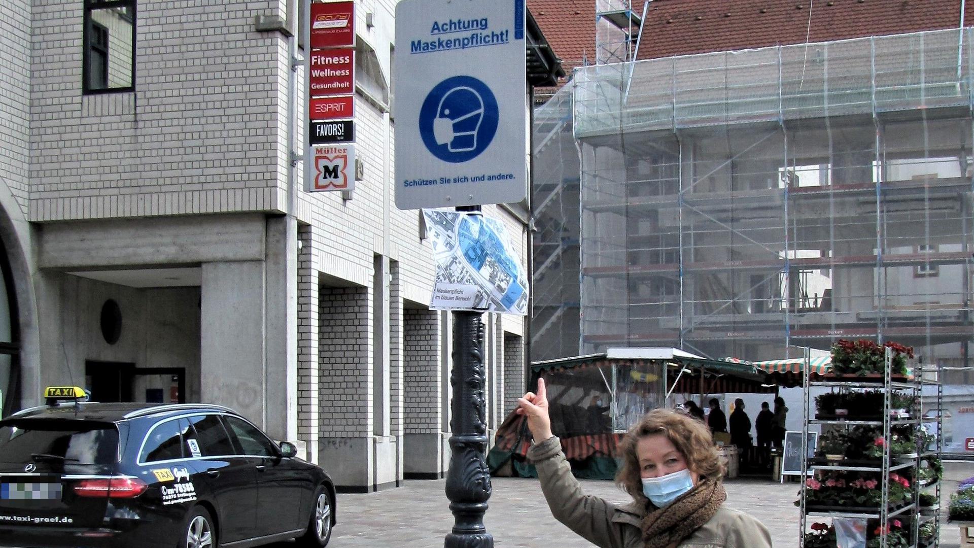 Ruth Lingenfelser steht vor einem Schild mit der Aufschrift „Achtung Maskenpflicht“ in Ettlingen, vor ihr auf dem Boden liegt eine getragene Corona-Maske.