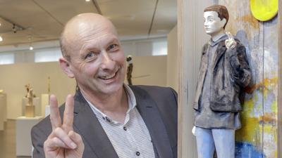 Bildhauer Rudi Bannwarth steht neben seinem Werk und lächelt in die Kamera. Mit den Händen formt er das Victory-Zeichen.