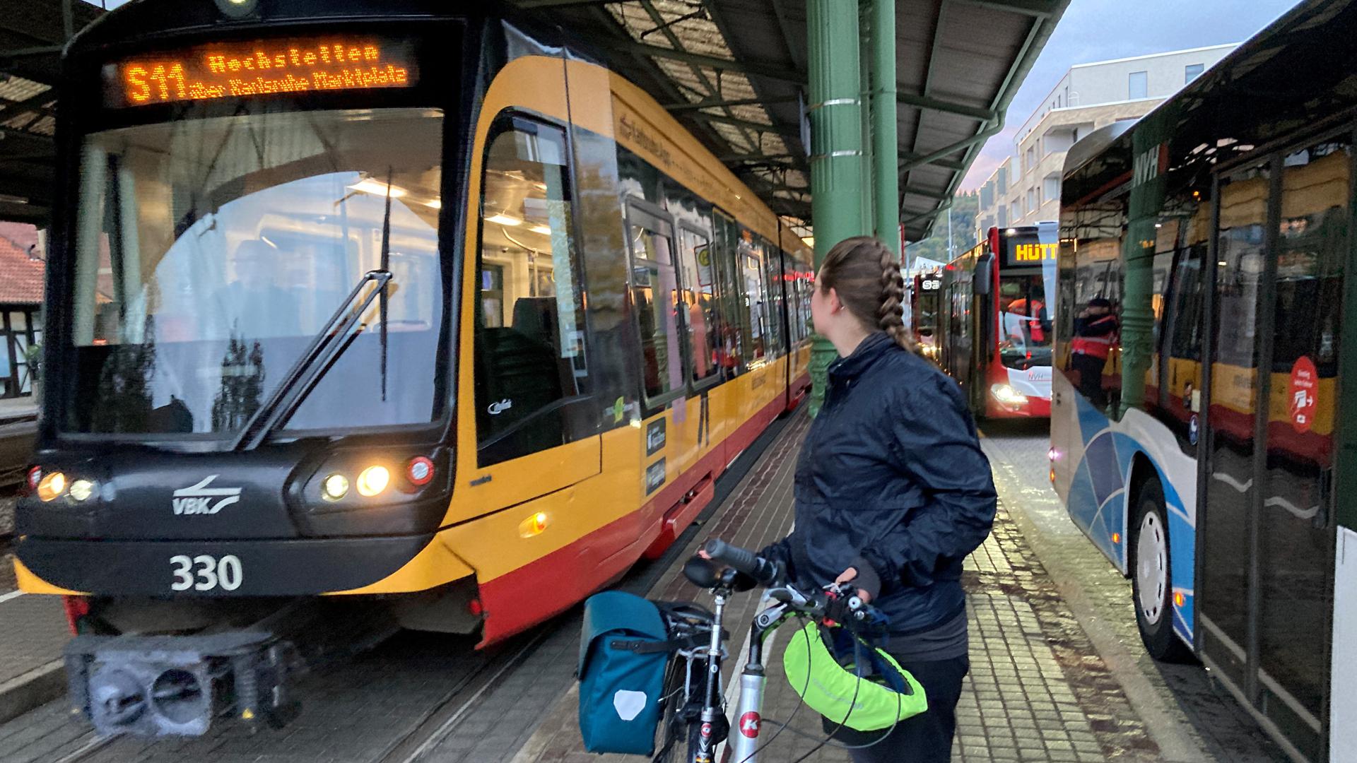 Eine Frau steht mit ihrem Fahrrad zwischen einer Stadtbahn und wartenden Bussen auf dem Bahnsteig.