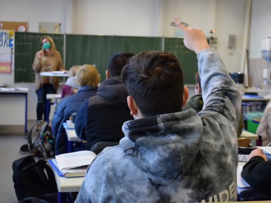 Schüler sitzen in einem Klassenraum der Schillerschule und schauen zur Lehrerin an der Tafel. Ein Schüler zeigt auf. 