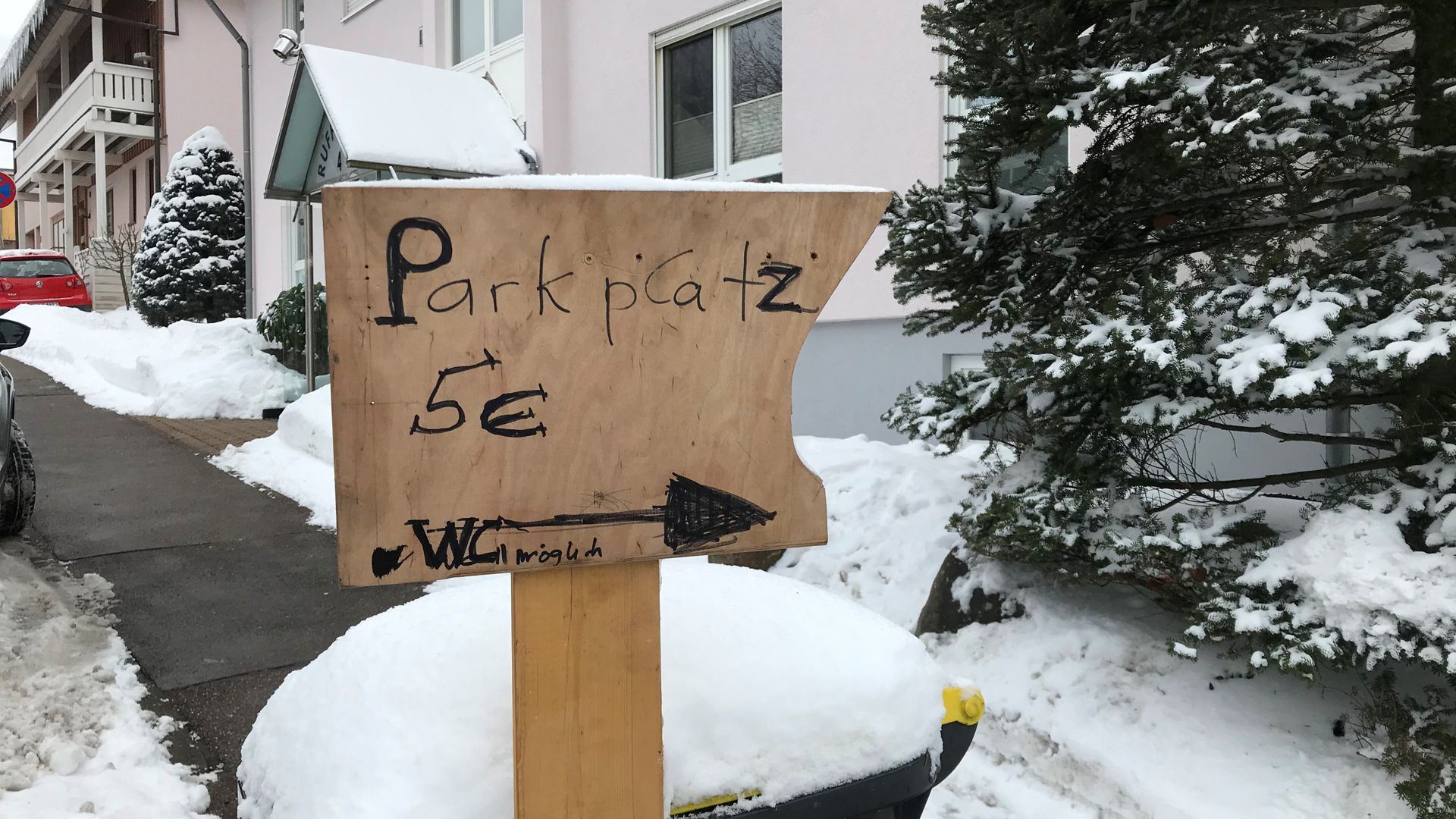 „Parkplatz 5 Euro, WC möglich“ steht auf einem Schild, das am Straßenrand in Dobel an einer mit Schnee bedeckten Mülltonne lehnt. Ein Pfeil zeigt nach rechts.