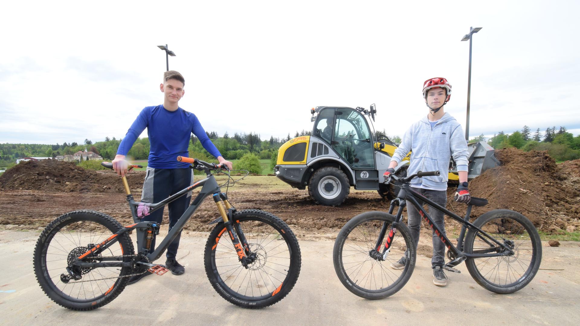 Maxim Ostrowski und Philip Sagawe stehen mit ihren Fahrrädern vor dem Gelände am Karlsbader Schulzentrum, auf dem der neuen Pumptrack entstehen soll. Im Hintergrund verteilen Bagger die Erde für den neuen Parcours.