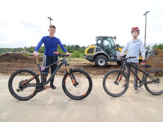 Maxim Ostrowski und Philip Sagawe stehen mit ihren Fahrrädern vor dem Gelände am Karlsbader Schulzentrum, auf dem der neuen Pumptrack entstehen soll. Im Hintergrund verteilen Bagger die Erde für den neuen Parcours.