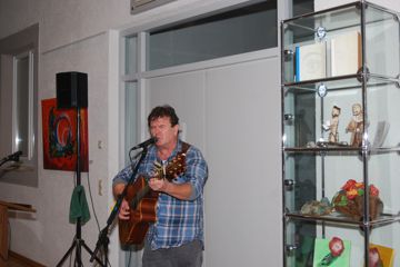 Ein Mann steht vor einem Mikrofon. Er singt und spielt Gitarre. Rechts neben ihm ist eine Vitrine mit Skulpturen darin.