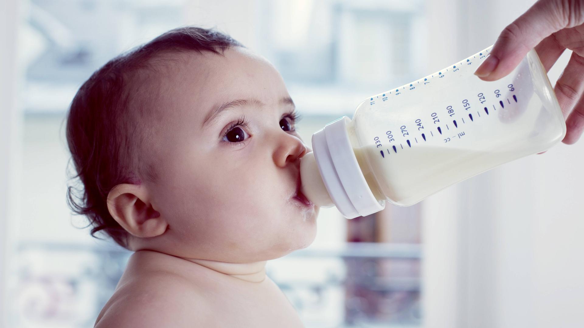 Ein sitzendes Kleinkind trinkt Milch aus einem Fläschchen.