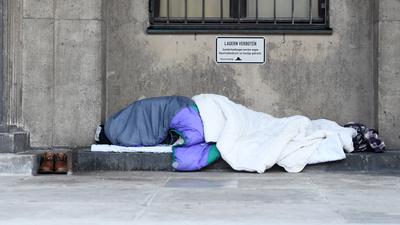 Ein Obdachloser schläft in Decken und Schlafsäcke gehüllt unter einem Verbotsschild mit der Aufschrift «Lagern verboten» an einem öffentlichen Gebäude.