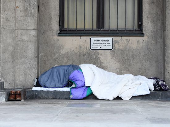 Ein Obdachloser schläft in Decken und Schlafsäcke gehüllt unter einem Verbotsschild mit der Aufschrift «Lagern verboten» an einem öffentlichen Gebäude.