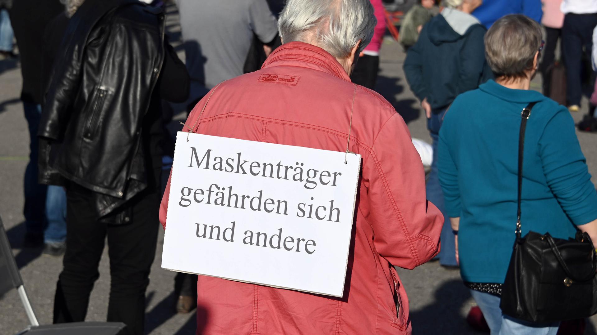 Ein Teilnehmer einer Karlsruher „Querdenken“-Demonstration gegen die Anti-Corona-Maßnahmen hat ein Schild umhängen, auf dem „Maskenträger gefährden sich und andere“ steht.