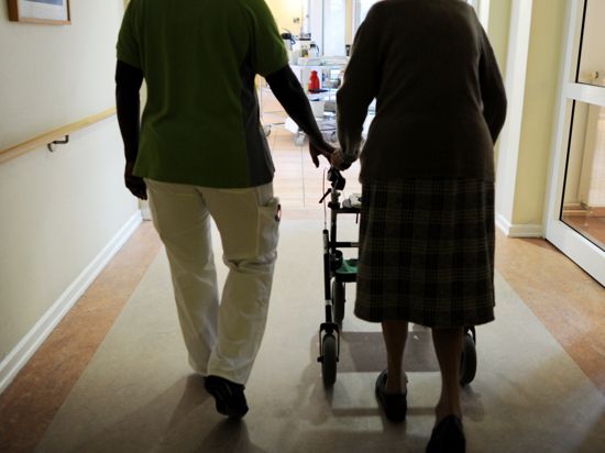 Eine Pflegehausbewohnerin und eine Pflegehelferin gehen mit einem Rollator einen Gang entlang. 
