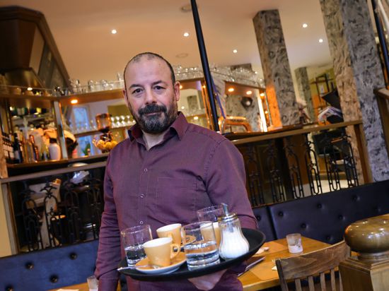 Alessandro Muccardo hält ein Tablett in der Hand, auf dem drei Espresso-Tassen, drei Wassergläser und ein Zuckerspender stehen. Im Hintergrund sind das Restaurant und die Theke zu sehen.