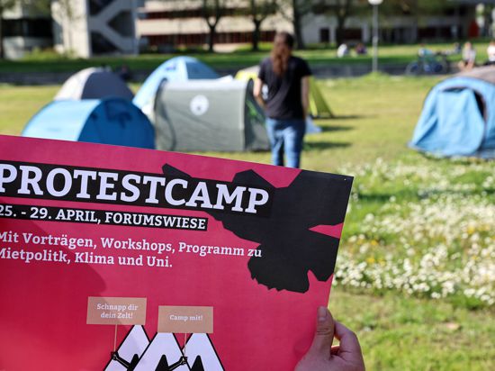 Jemand hält einen pinken Info-Flyer in die Höhe, im Hintergrund sieht man Zelte des Protestcams