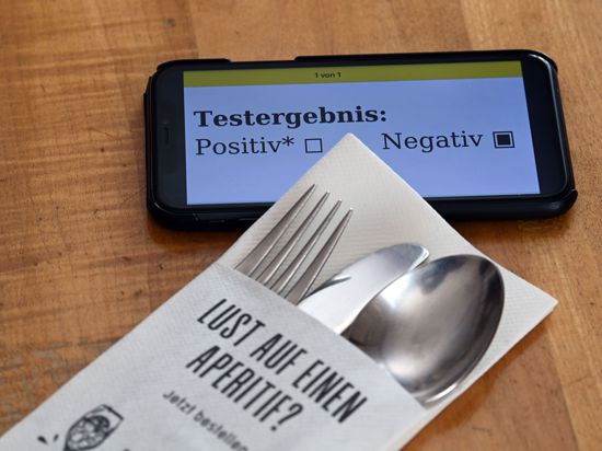 Ein Serviettenetui mit Gabel, Messer und Löffel und dem Aufdruck «Lust auf einen Aperitif?» liegt in einem Restaurant neben einem Smartphone, dessen Display ein negatives Corona-Testergebnis zeigt.