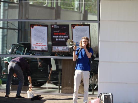 Aktion der Letzten Generation in Karlsruhe: Ein Demonstrant steht mit einem Megafon vor einer Glaswand. An dieser werden Plakate aufgeklebt.