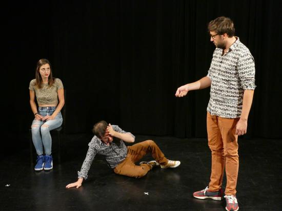 Drei Personen auf einer dunklen Theaterbühne, links eine sitzende Frau, die die Hände in den Schoß legt, in der Mitte liegt ein Mann am Boden, der Mann rechts von ihm schaut auf ihn herab.