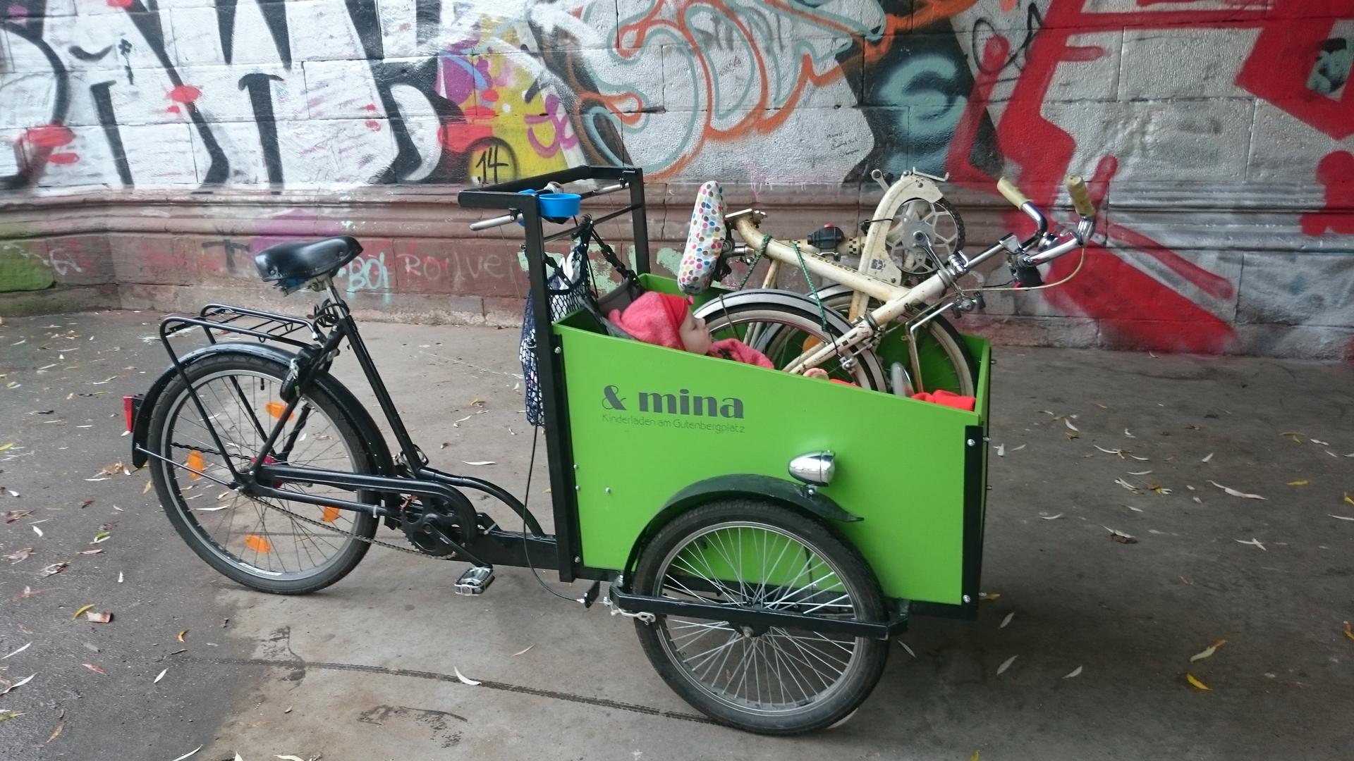 In der froschgrünen Transportbox eines Lastenrads sitzt ein zufriedenes kleines Kind neben einem zusammengeklappten Faltrad.