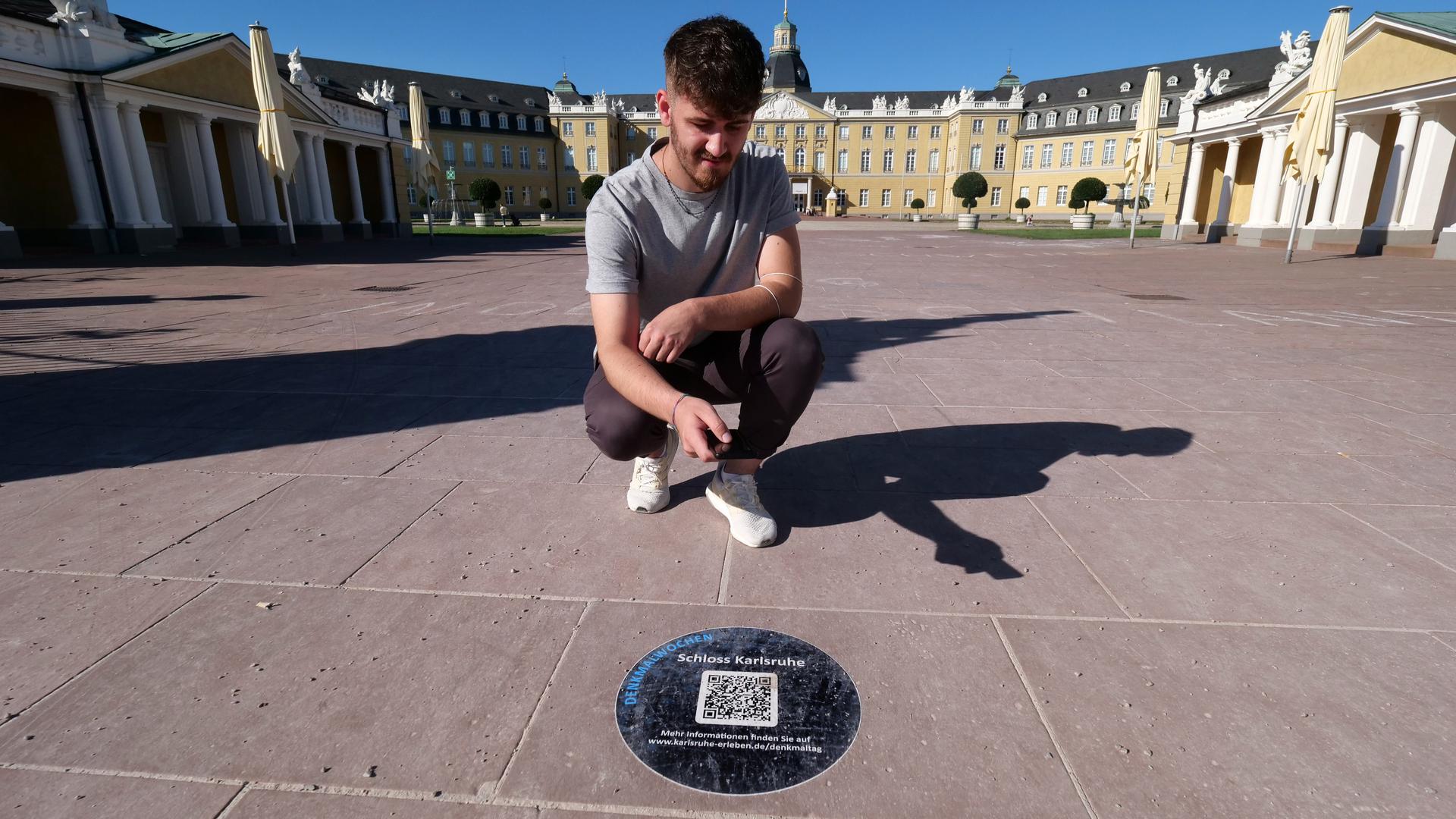 Ein junger Mann kniet vor dem Karlsruher Schloss und scannt mit seinem Handy einen QR-Code ein.