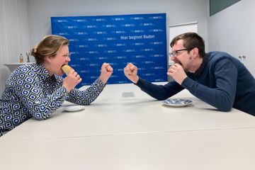 Redakteurin Judith Midinet-Horst (links) und Redakteur Arne Oest (rechts) sitzen sich an einem Tisch gegenüber. Sie beißt in ein süßes Teilchen, er in einen Burger. 