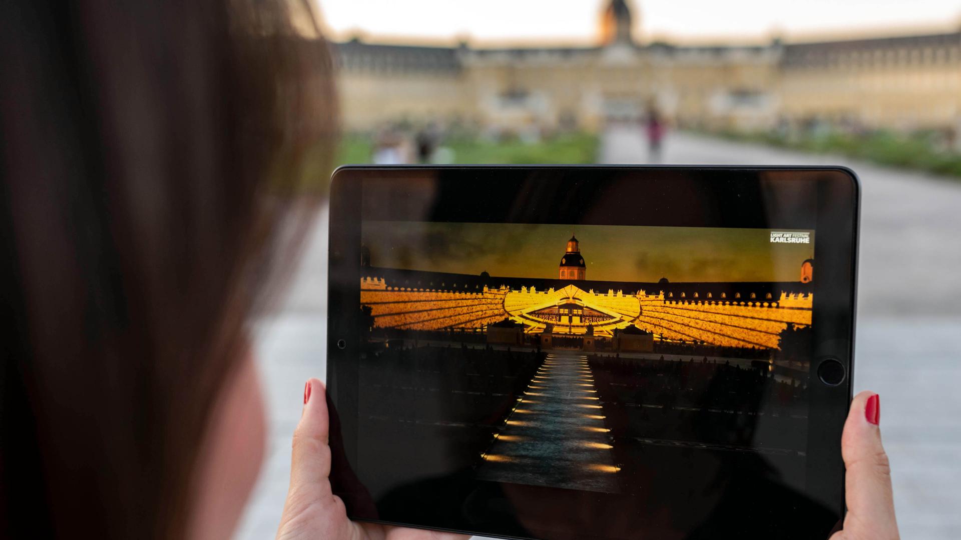 Vor dem Schloss Karlsruhe betrachtet eine Person auf einem Tablet einen Teil der Schlosslichtspiele, die 2020 wegen der Corona-Pandemie virtuell im Internet stattfinden mussten.
