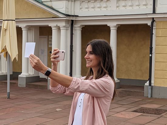 Eine junge Frau hebt vor dem Karlsruher Schloss ein ausgedrucktes Bild hoch und macht ein Foto davon.