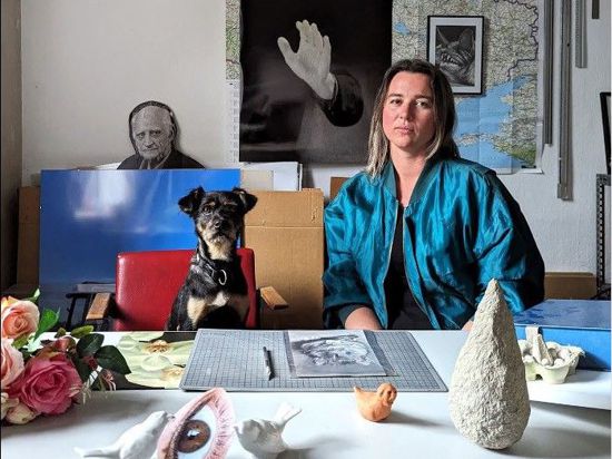Die Künstlerin Katharina Baumann an einem Tisch mit Kunstwerken neben einem Hund.