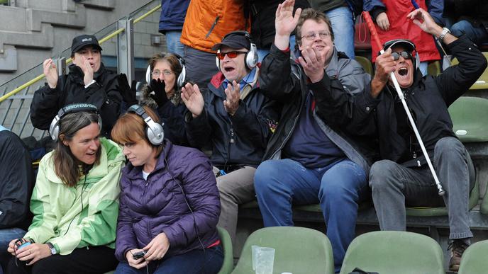 Fußball-Fans im Stadion mit Kopfhörern und Blindenstock.