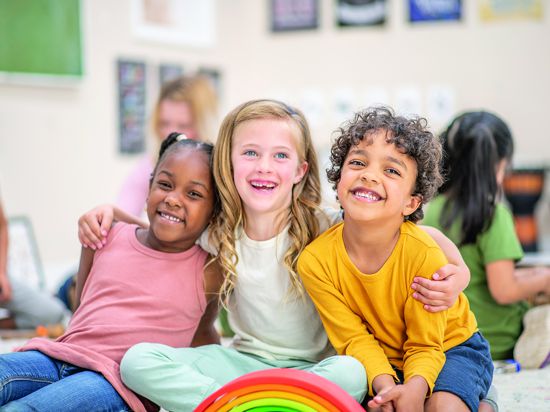 A multi-ethnic group of daycare children sit close together on the floor, with their legs crossed and their arms around one another, as they pose for a portrait. They are smiling and enjoying their time together.