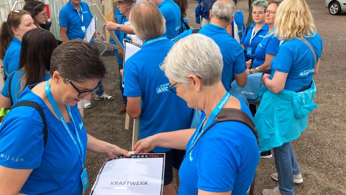 Volunteers in leuchtend blauen Shirts bereiten beim Kraftwerk-Open-Air-Konzert am Schloss Infoschilder mit Geländeskizzen vor.