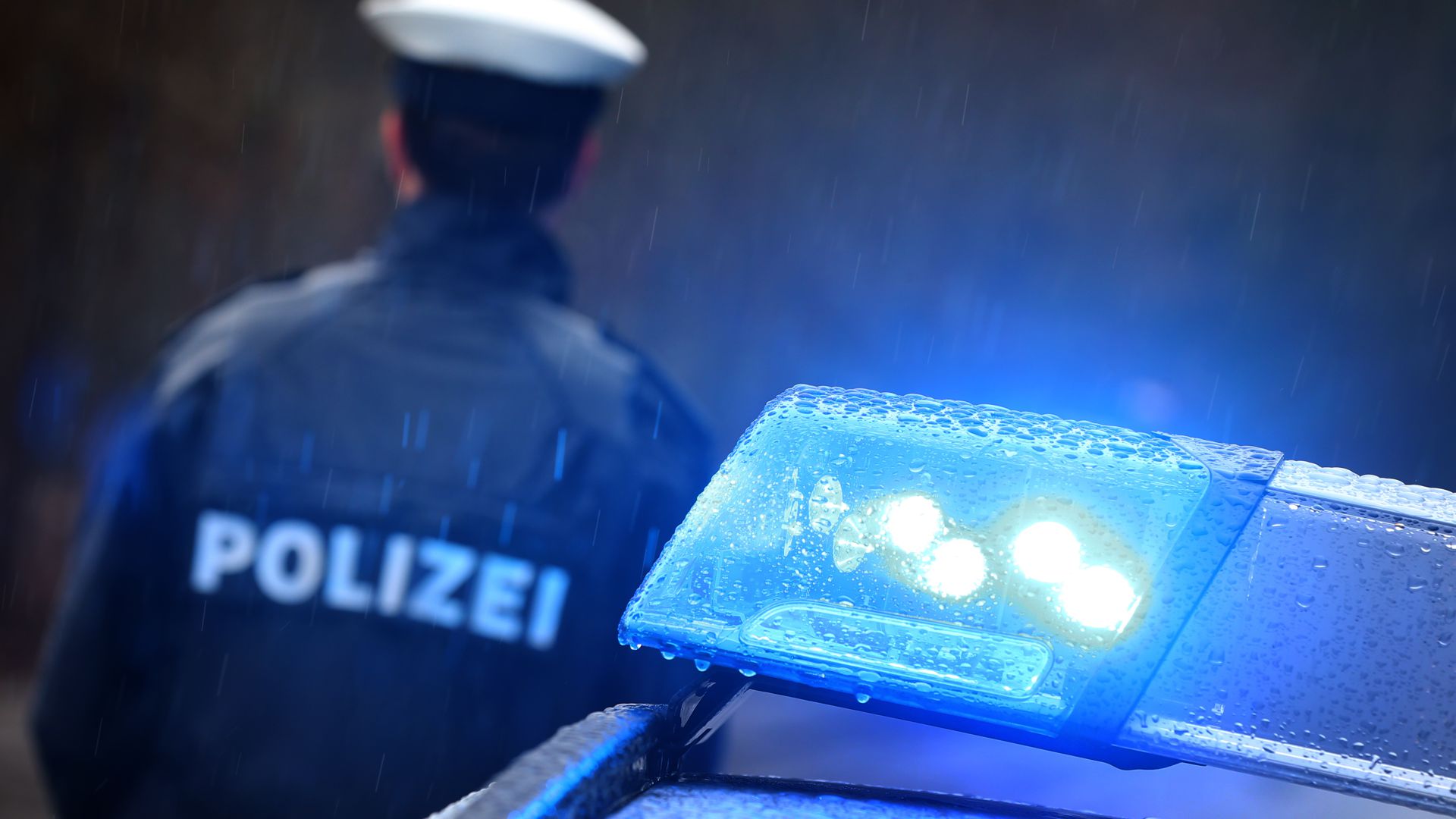  Ein Polizist steht im Regen vor einem Streifenwagen, dessen Blaulicht aktiviert ist. 