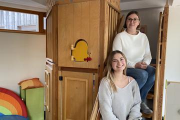 Die PiA-Auszubildende Sophie Schenk und Kita-Leiterin Laura Schehrer sitzen auf der Treppe eines Spielzeughauses in der städtischen Kindertageseinrichtung Sybelstraße.