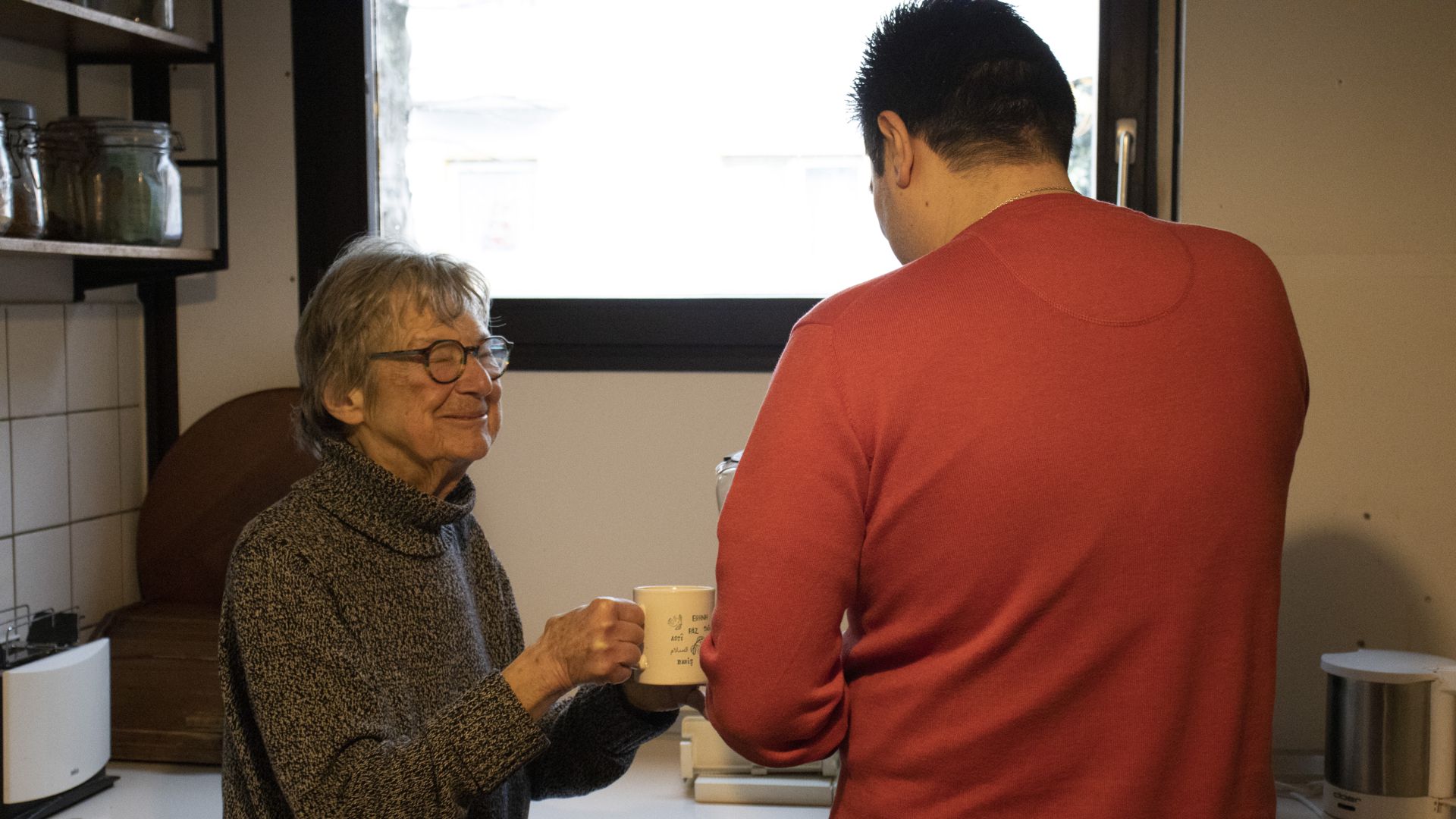 Links steht eine ältere Dame, hebt einen Kaffeebecher. Rechts schenkt ein junger Mann der Dame Kaffee ein.