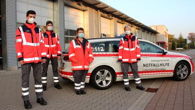 Vier maskierte Helfer in Schutzkleidung stehen vor Einsatzwagen der Notfallhilfe Linkenheim-Hochstetten