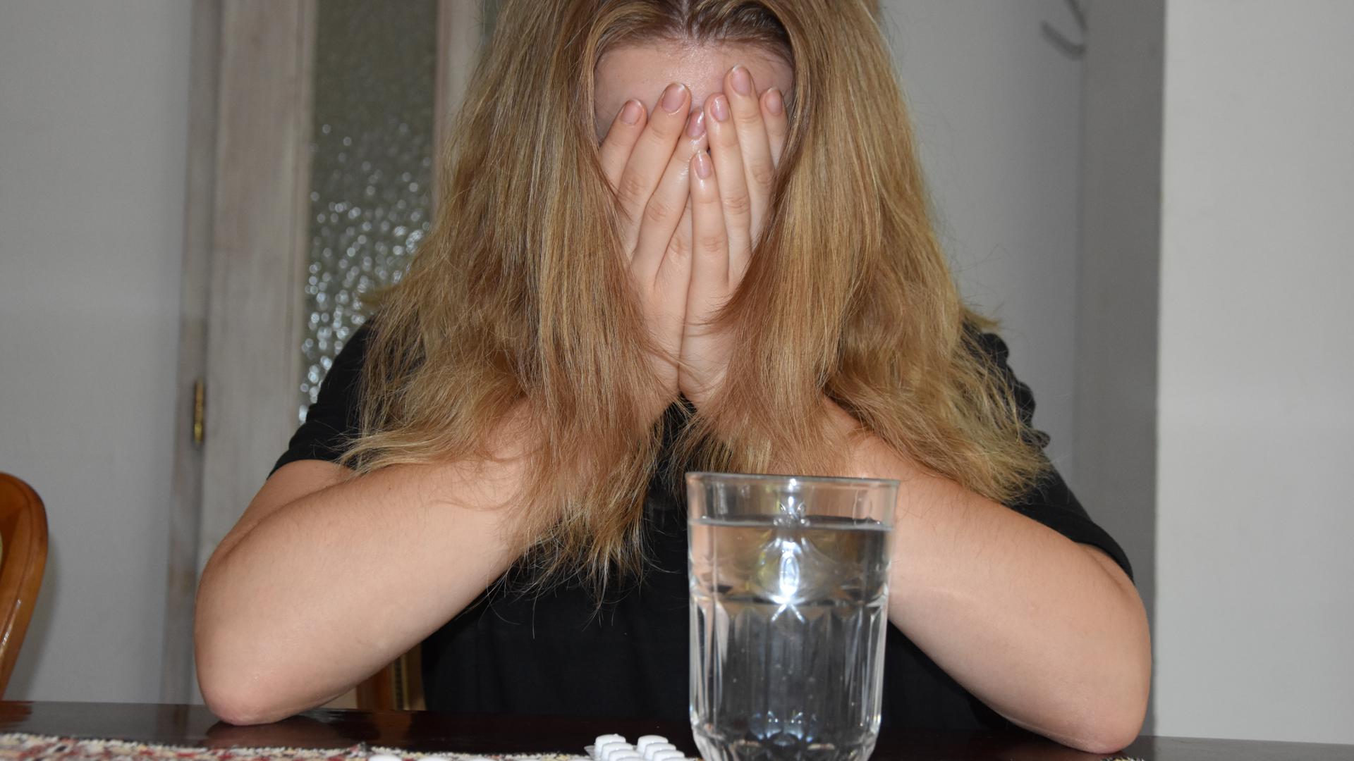 Frau bedeckt das Gesicht mit den Händen, sitzt am Tisch mit Tabletten und einem Wasserglas