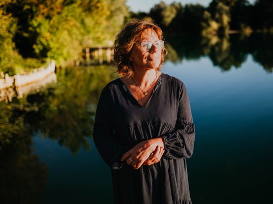 Eine Frau mit Brille steht vor einem See. 