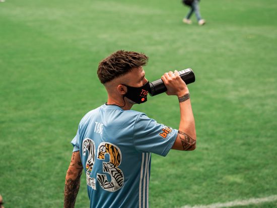Ein Mann mit blauem Trikot und Maske trinkt am Rand eines Fußballfelds aus einer Flasche. 