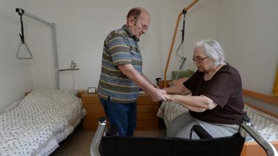 Ein älterer Mann pflegt seine Frau und hilft ihr, aus einem Krankenbett aufzustehen. 