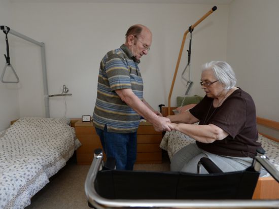 Ein älterer Mann pflegt seine Frau und hilft ihr, aus einem Krankenbett aufzustehen. 