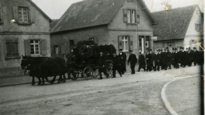 Eine Schwarz-weiß-Aufnahme zeigt einen Begräbniszug in Eggenstein-Leopoldshafen vor 1945, voran fährt eine schwarze Kutsche mit dem Sarg. 