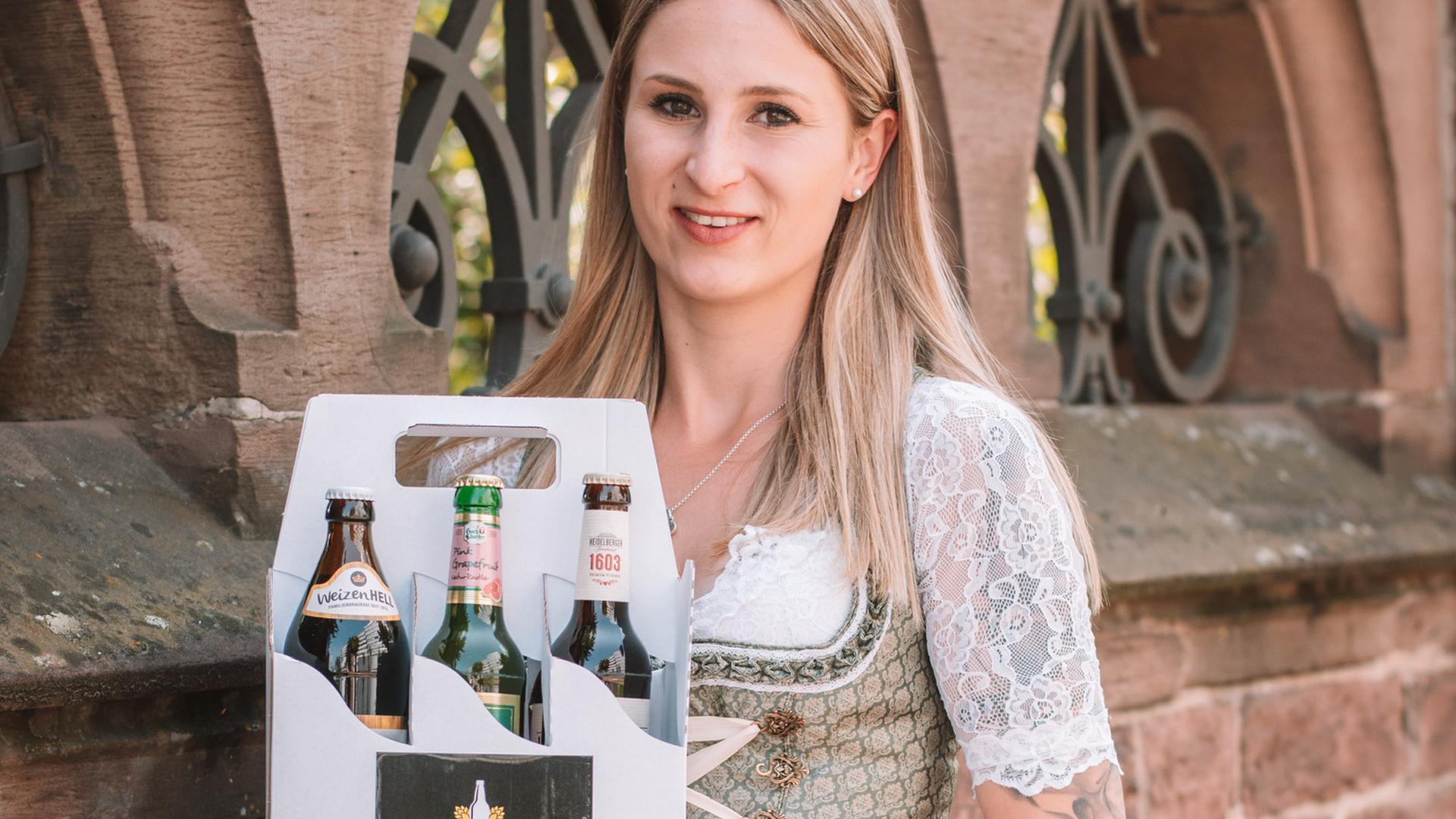 Junge Frau mit langen Haaren im Dirndkleid hält einen Karton mit verschiedenen Biersorten hoch.