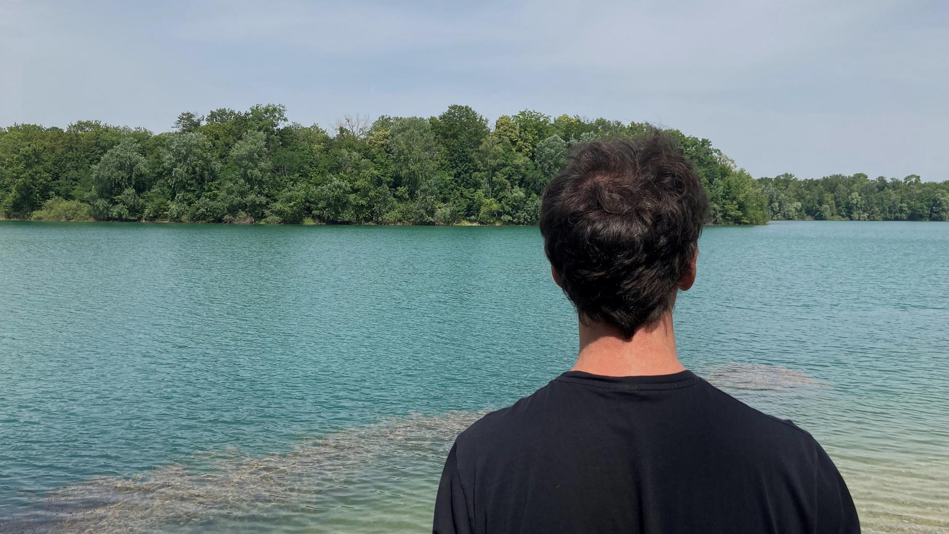 Ein dunkelhaariger Mann blickt auf einen See. 