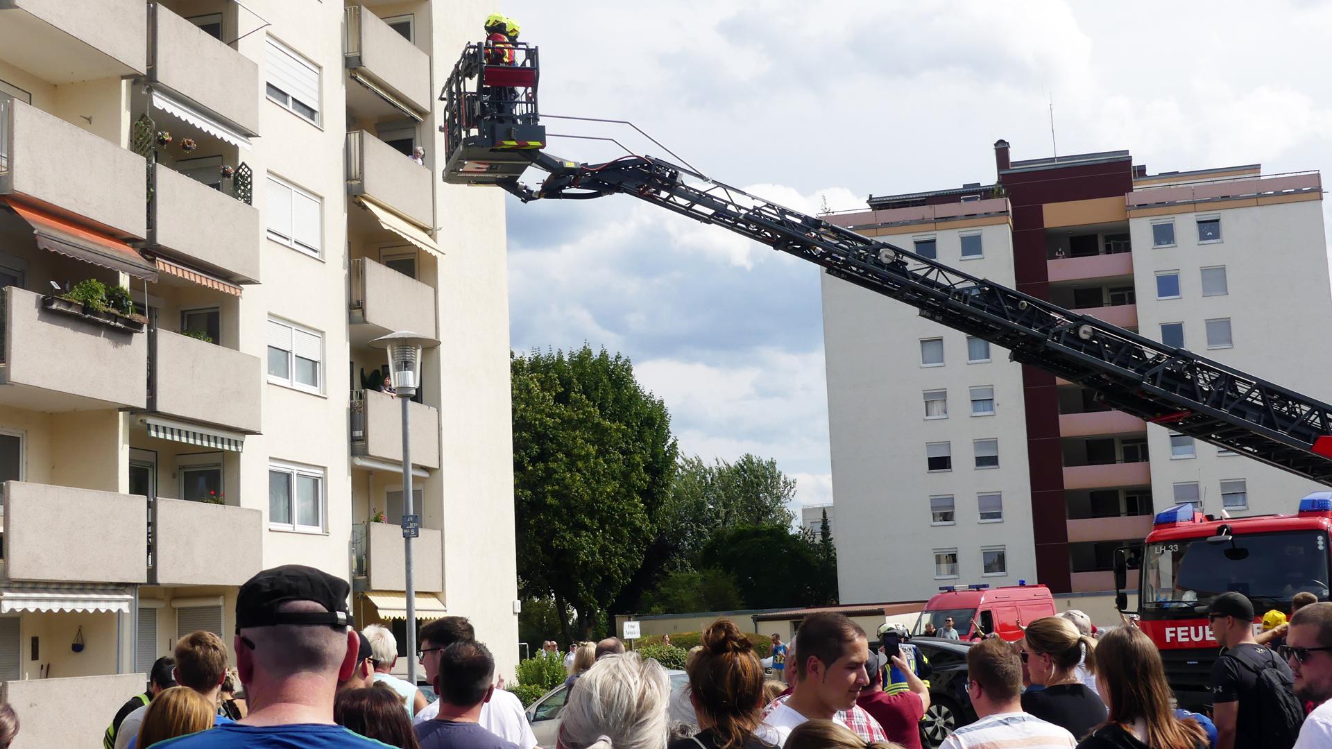Viele Menschen schauen der Feuerwehr zu. Eine Drehleiter befördert Retter zu einem Balkon an einem Wohnblock.  